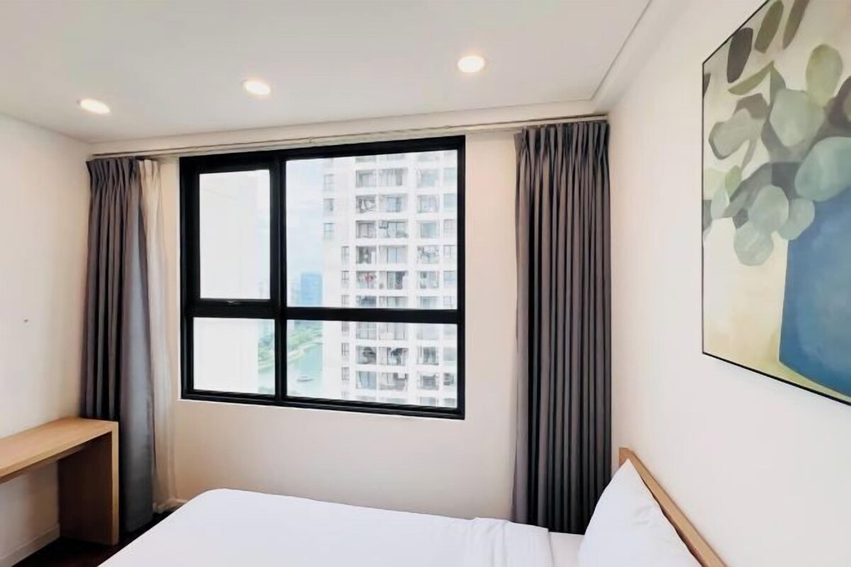 A hotel room with a bed, a desk, a window with curtains, and a painting on the wall.