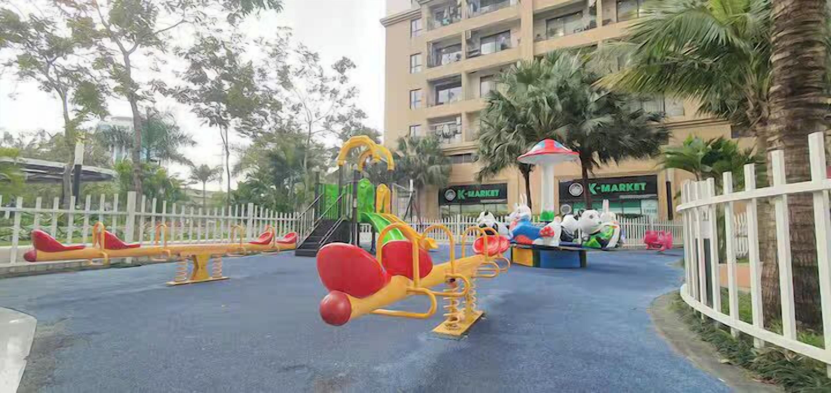 Ein Spielplatz im Freien mit Wippen und einer Kletterkonstruktion, umgeben von einem weißen Zaun und Palmen.