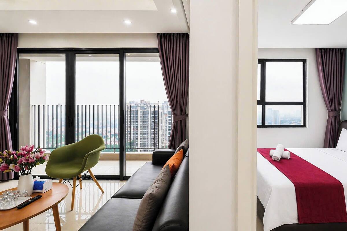 A hotel room with a balcony, a bed, a sofa, a table with a vase of flowers, and a green chair.