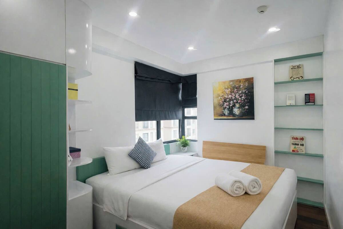 A hotel room with a bed, a green wardrobe, a window with a view, a painting on the wall, and a shelf with items.