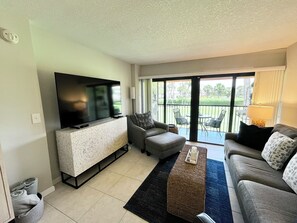 A living room with a flat-screen TV, grey sofas, a coffee table, and a balcony with outdoor furniture.