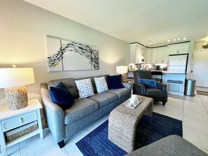 A modern living room with a grey sofa, a coffee table, a lamp, and a kitchen in the background. - OV 926 Golf Course View Condo - Welcome to Paradise (Fort Pierce)