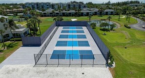 Un court de tennis entouré d’un complexe hôtelier comprenant plusieurs bâtiments, avec un terrain de golf en arrière-plan.