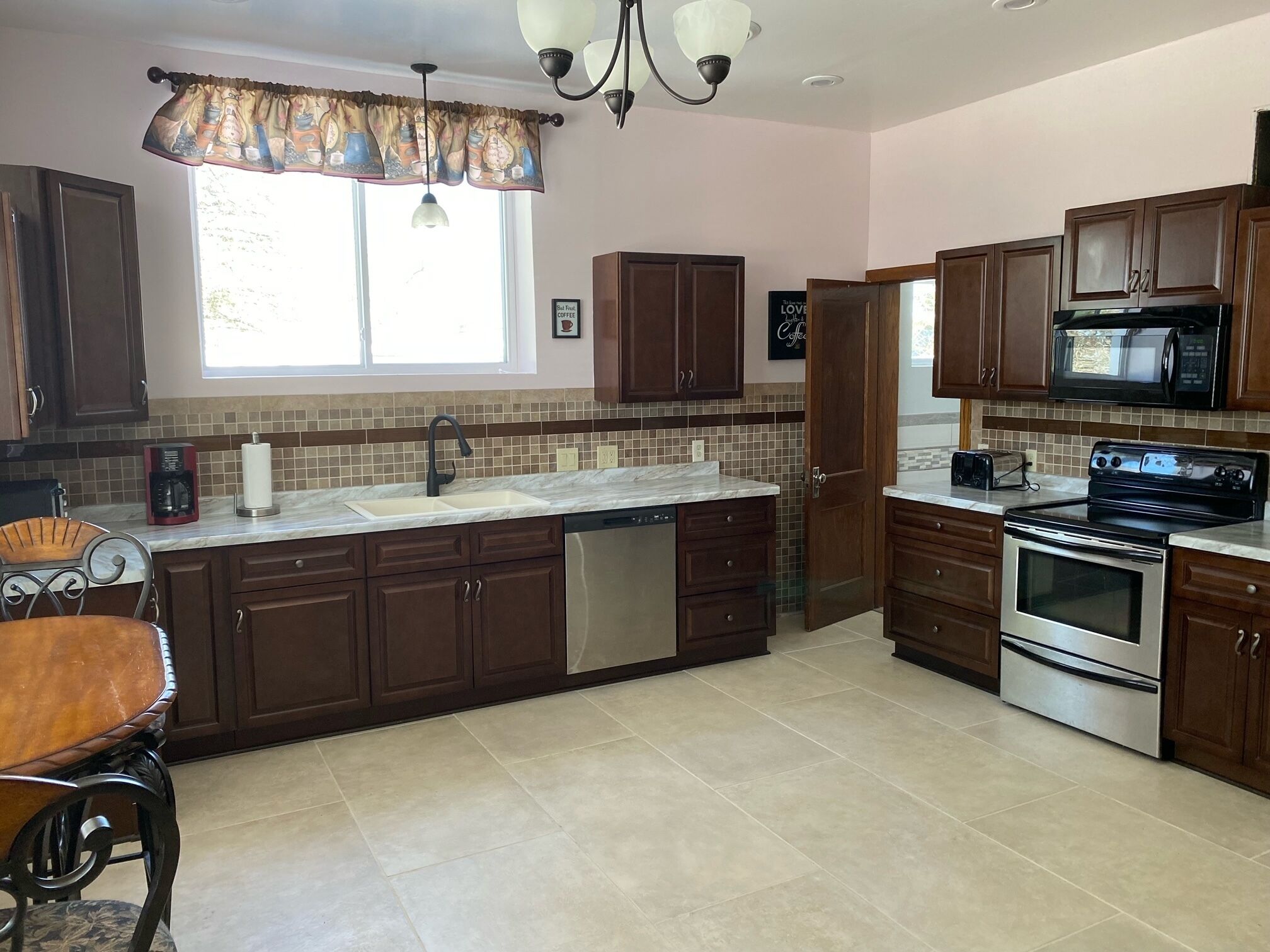 A kitchen with dark wooden cabinets, a stainless steel dishwasher, a double oven, a microwave, and a large window with a curtain.