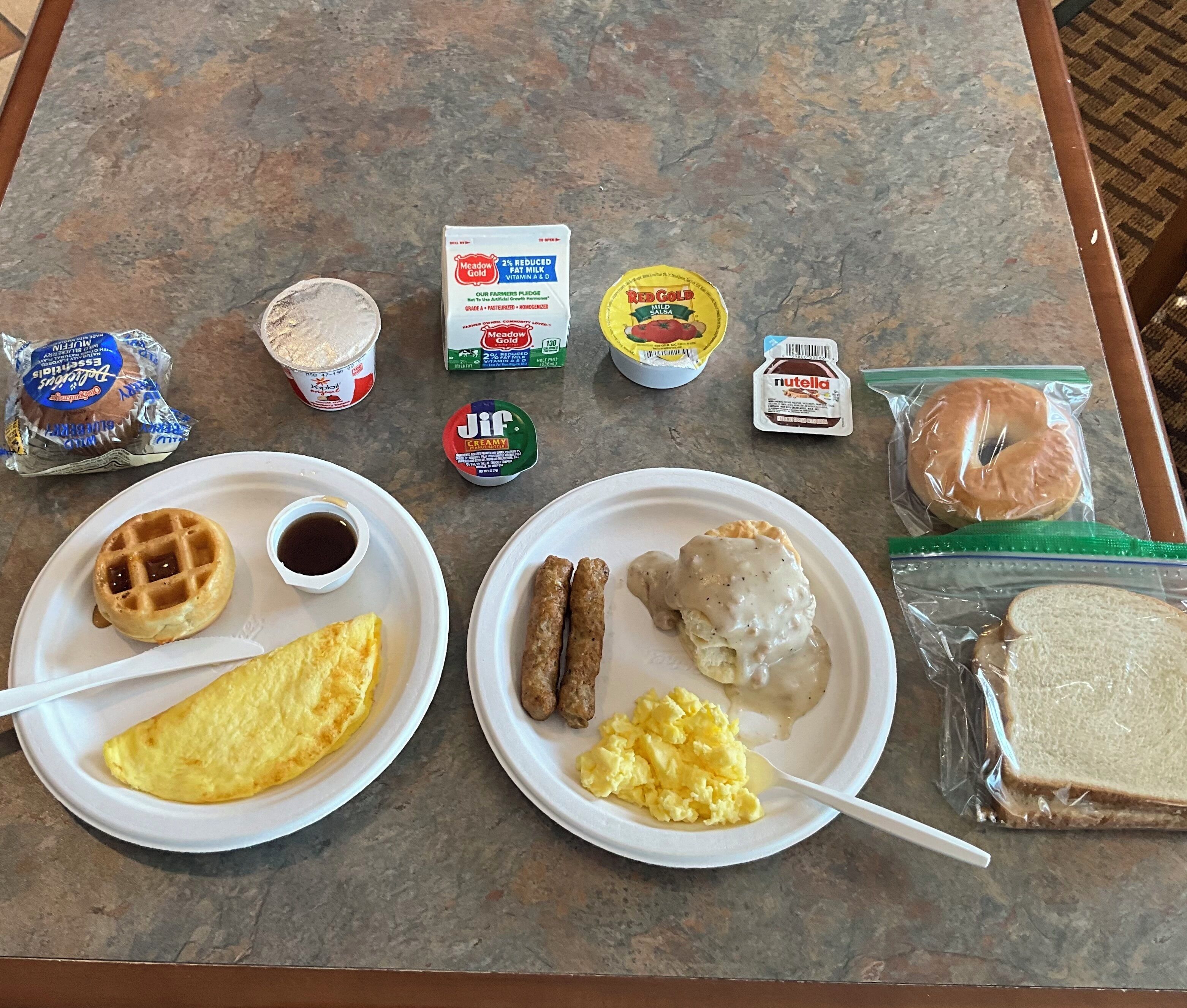 A breakfast spread on a table including waffles, scrambled eggs, sausages, biscuits, and various spreads.