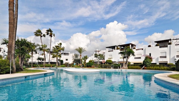 Una piscina con agua azul cristalina, rodeada de palmeras y edificios blancos con balcones.