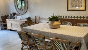 Un comedor con una mesa redonda cubierta por un mantel estampado, rodeado por cuatro sillas de mimbre con cojines de diseño en zigzag. Una planta en maceta se coloca sobre la mesa, y un espejo decorativo cuelga en la pared encima.