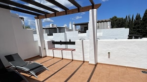 Un patio techado al aire libre con una silla de salón y una mesita pequeña, rodeado de paredes blancas y un suelo de baldosas.