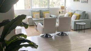 A modern living room with a sofa set, two armchairs, a coffee table, and a side table. There is a large window with a view of trees and a lamp on the side table.