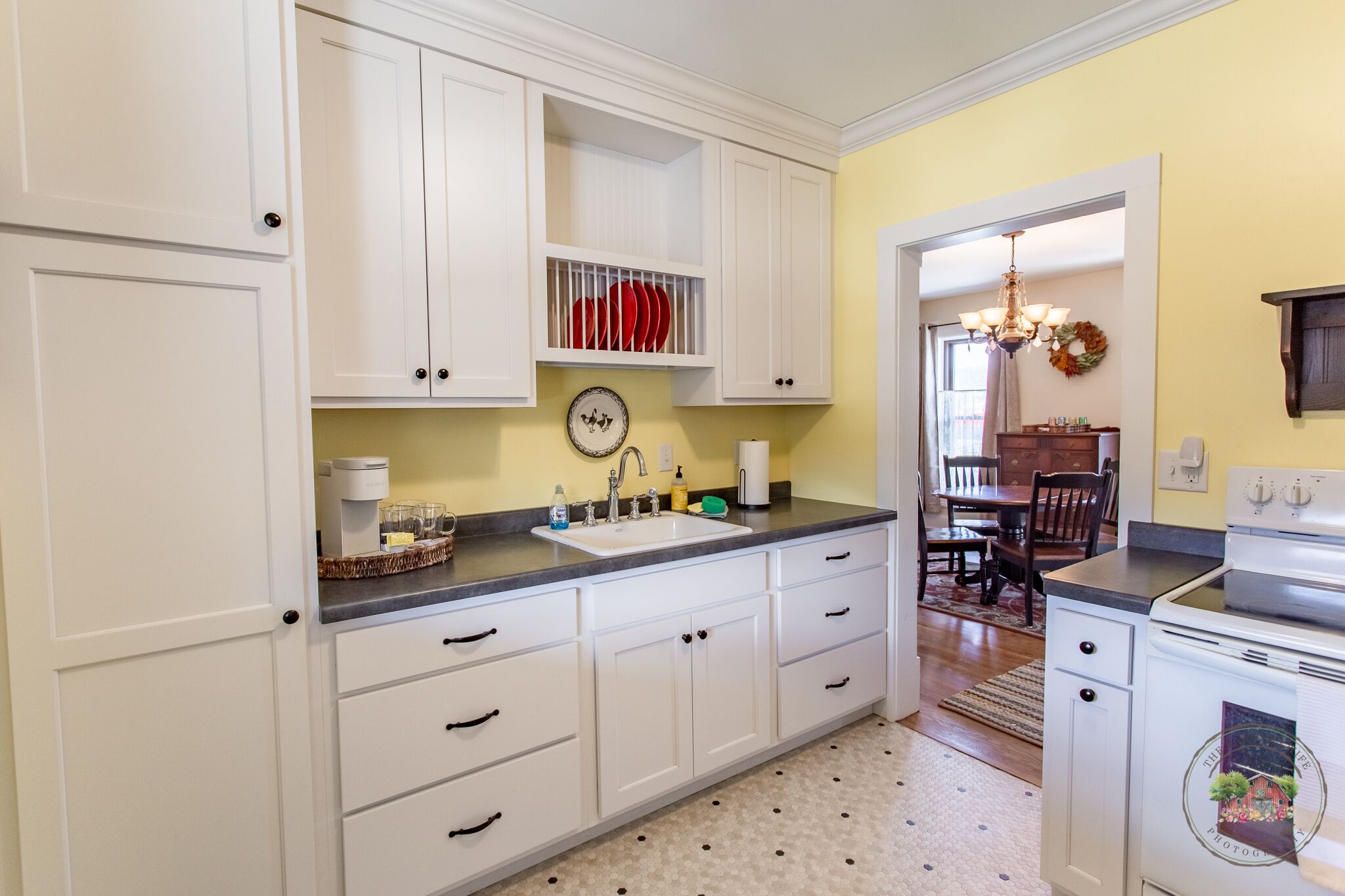 Une cuisine avec des meubles blancs, un évier et une cuisinière. On aperçoit un coin repas avec une table et des chaises à travers une porte ouverte.