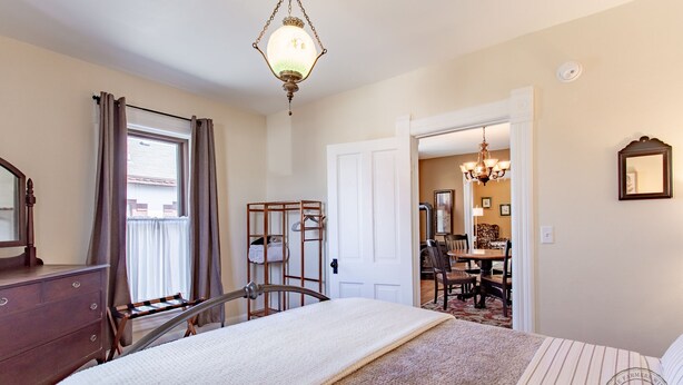 A bedroom with a bed, a dresser, a mirror, a window with curtains, a chandelier, and a dining area visible through an open door. - Charming Downtown Two Bedroom w/ Fireplace (New Glarus)