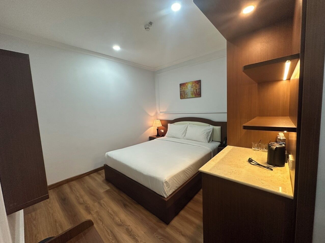 Un dormitorio con cama, mesita de noche, lámpara y un estante con objetos.