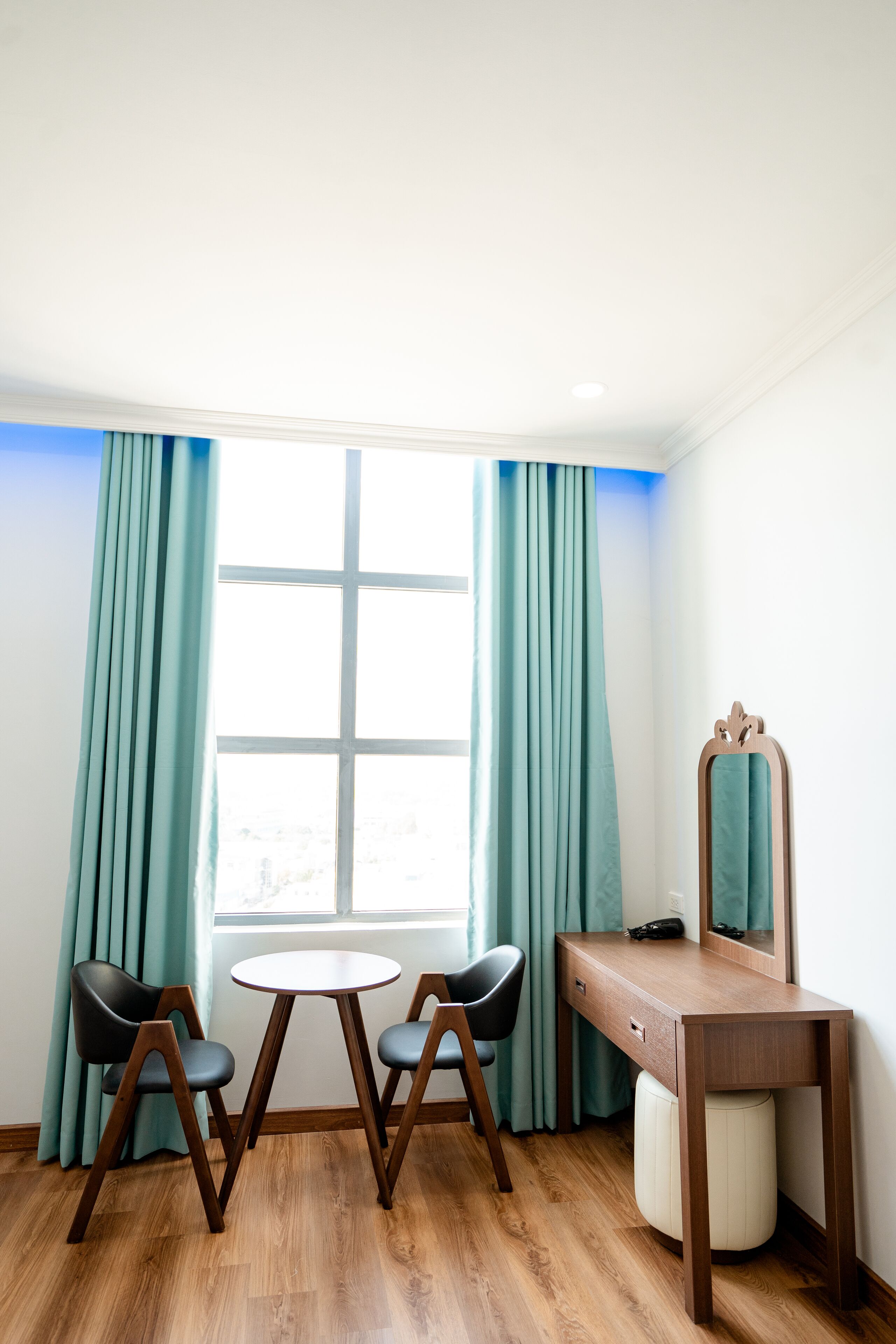 A room with a wooden desk, two chairs, a round table, a mirror, and teal curtains.
