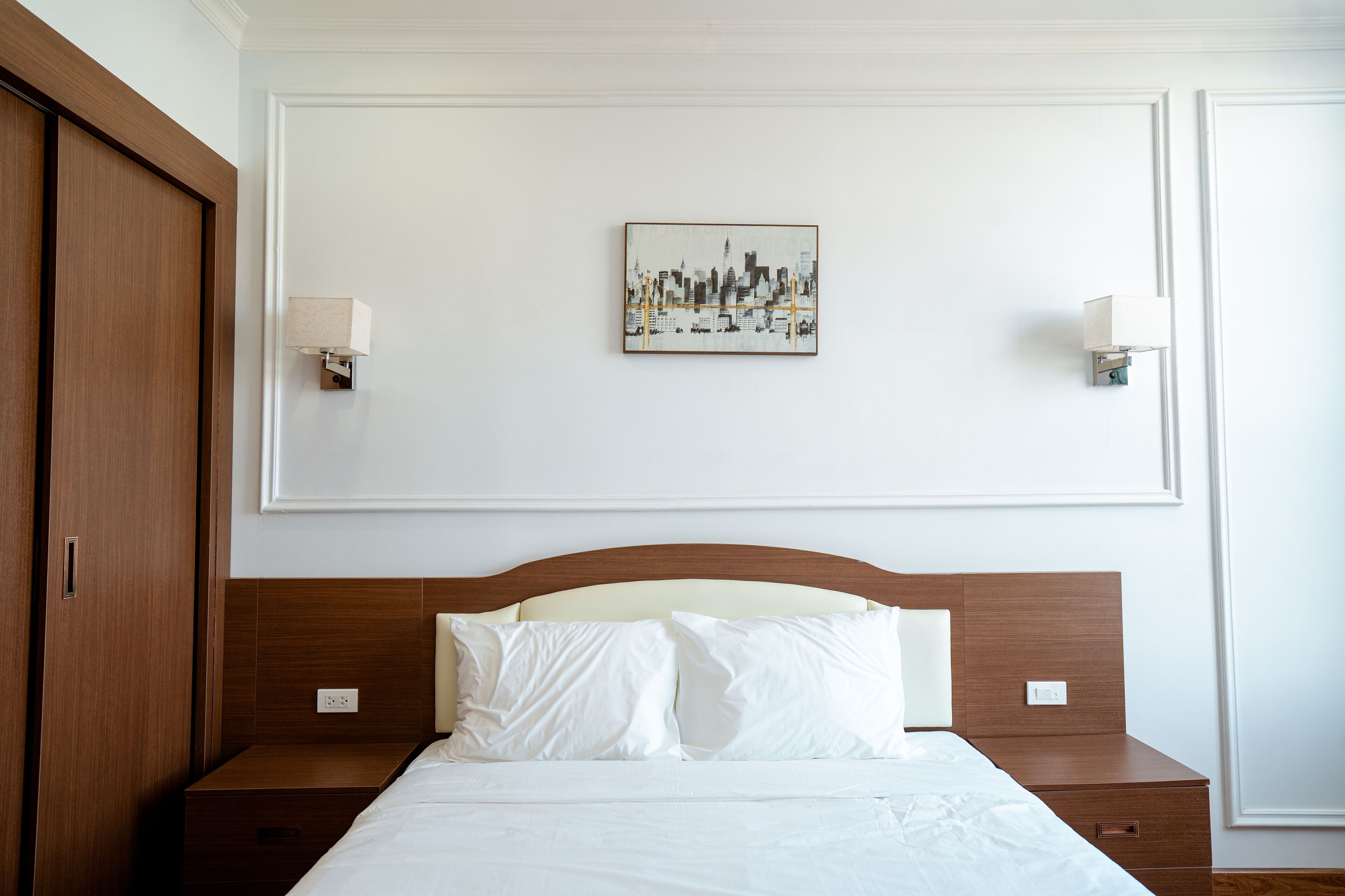 Camera d'hotel con una testiera in legno, una coperta bianca, due comodini e un'opera d'arte con una veduta sulla città.