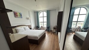 A modern hotel room with a large bed, a desk, and a chair. There are two windows with curtains, and a painting on the wall. - Ngoc Thu Palace (Can Tho)
