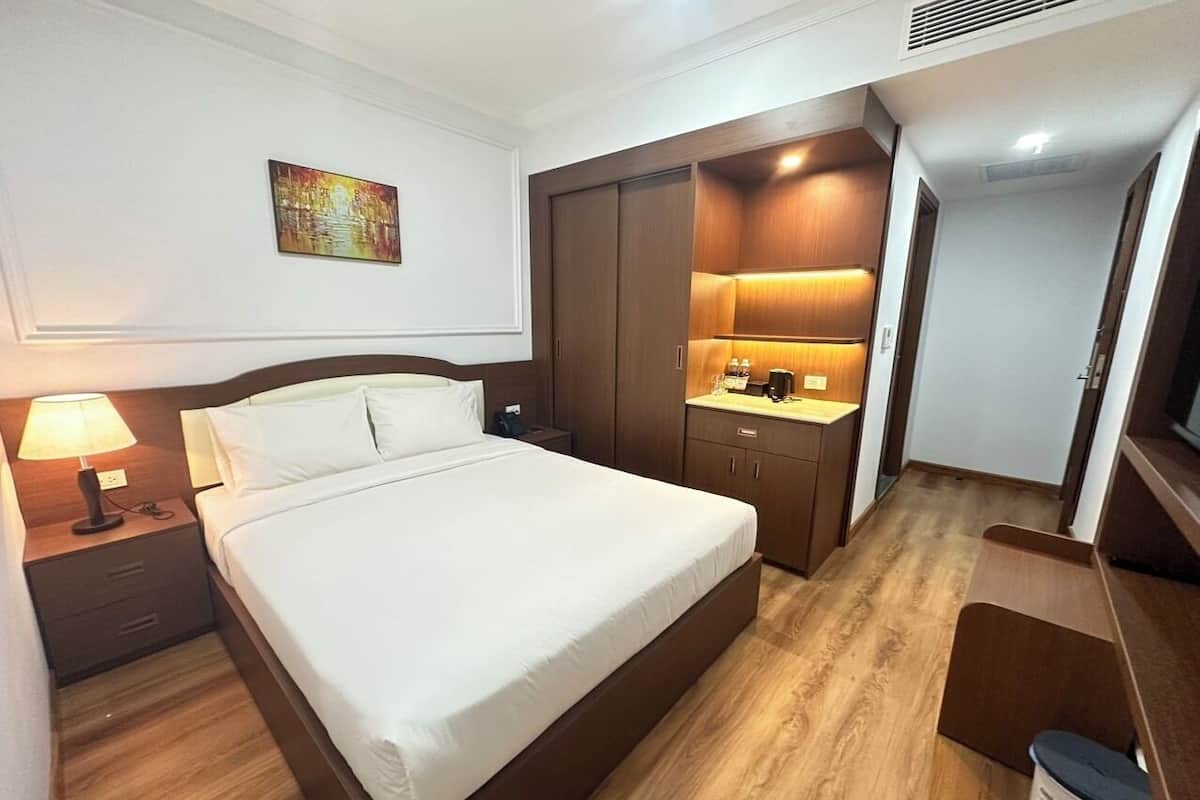 A hotel room with a large bed, a nightstand with a lamp, a wardrobe, a small desk with a lamp, and a television.