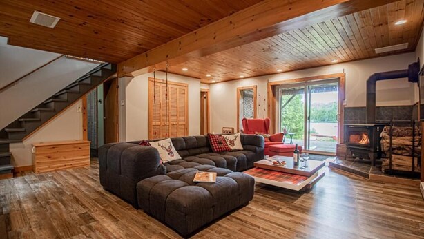 A spacious living room with a wooden floor, a sectional sofa, a red armchair, and a fireplace. - Peaceful Chalet on Pristine Lac de l'Abies in Labelle, QC. Dogs Welcome! (Labelle)