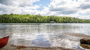 A lake with a forested shoreline and a boat on the sand. - Peaceful Chalet on Pristine Lac de l'Abies in Labelle, QC. Dogs Welcome! (Labelle)