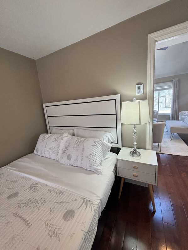A neatly arranged bedroom with a bed, bedside table, lamp, and a view of the adjacent room. - Home Away from Home! (Worcester)