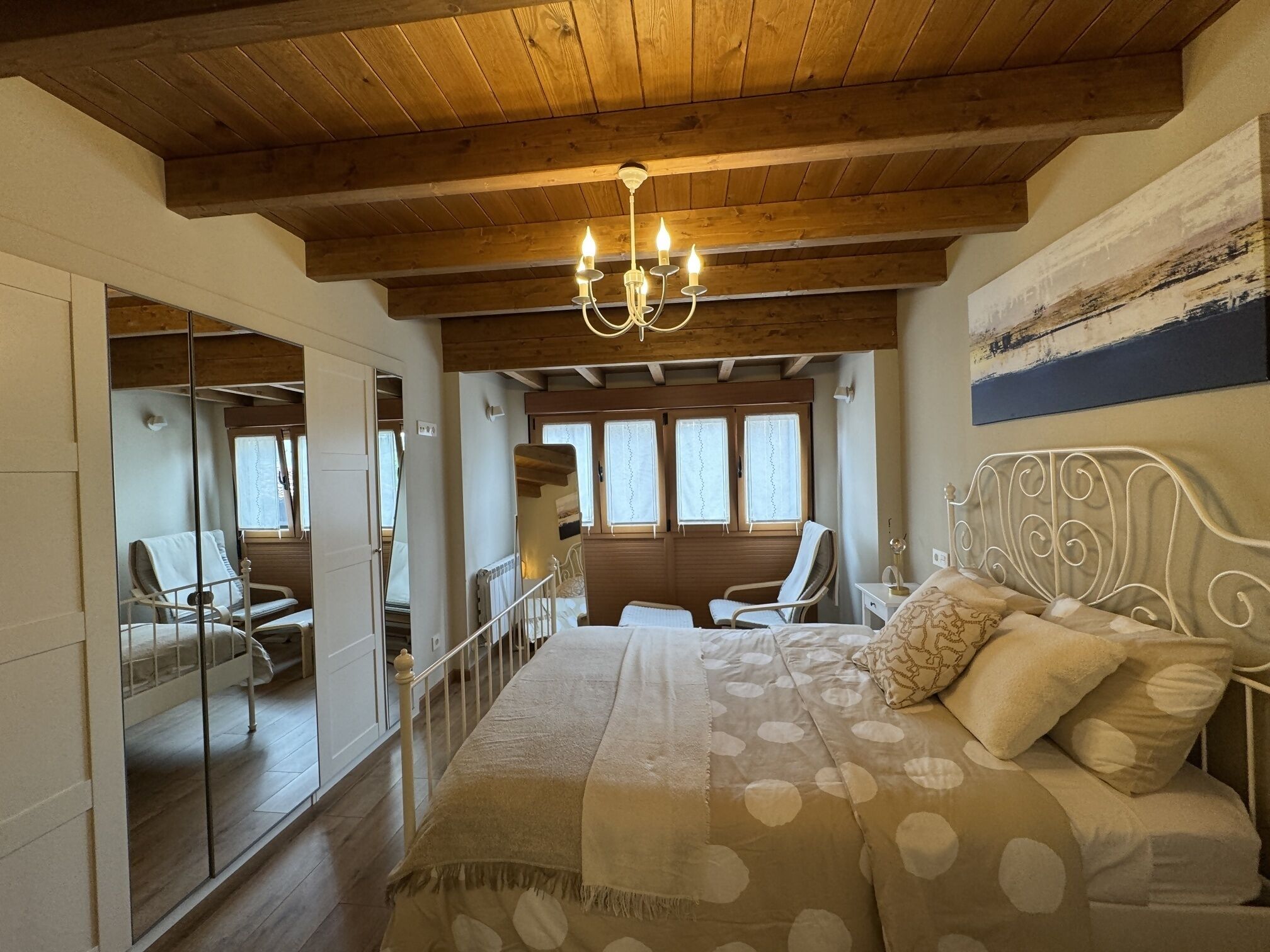 A bedroom with a wooden ceiling, a bed with a patterned bedspread, a chandelier, and a view of a balcony with chairs.