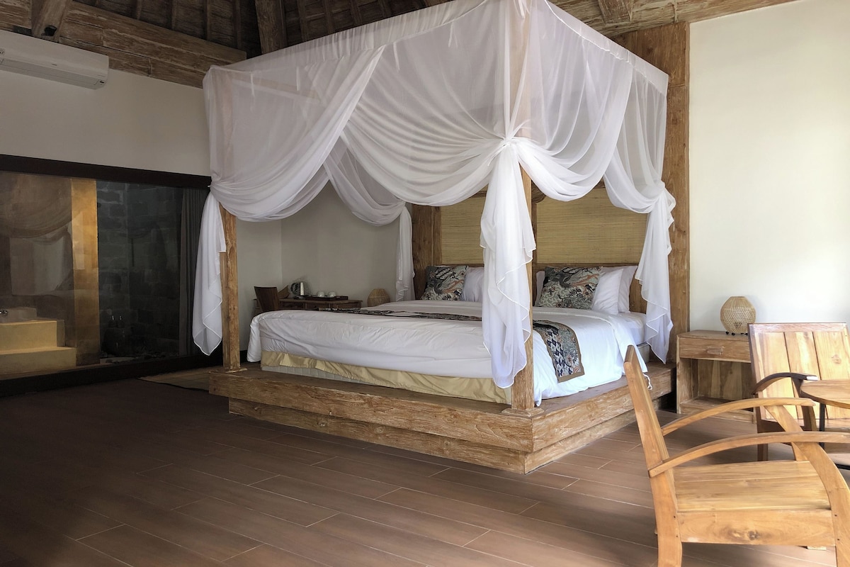 A four-poster canopy bed with a wooden frame and white bedding, a wooden nightstand with a lamp, and a wooden chair.