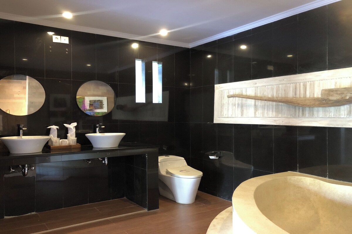 A modern bathroom with black tiles, two sinks, a toilet, and a bathtub.