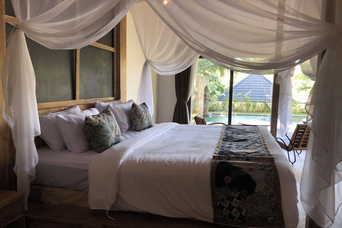A bedroom with a large bed, white bedding, and a decorative throw. There is a window with curtains, a view of a pool outside, and a wooden headboard.