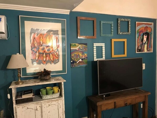 Ein Zimmer mit türkisfarbenen Wänden, einem Flachbildfernseher auf einem Holzbodenständchen, einer Lampe und verschiedenen gerahmten Kunstwerken.