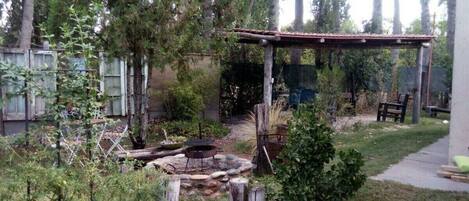 A backyard with a wooden pergola, a fire pit, and a small garden area with various plants and a bench.