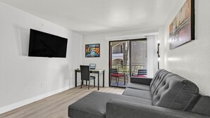 Ein modernes Wohnzimmer mit grauem Sofa, einem flachbildfernseher an der Wand, einem kleinen Tisch mit Stuhl und einem Balkon mit roten Stühlen.