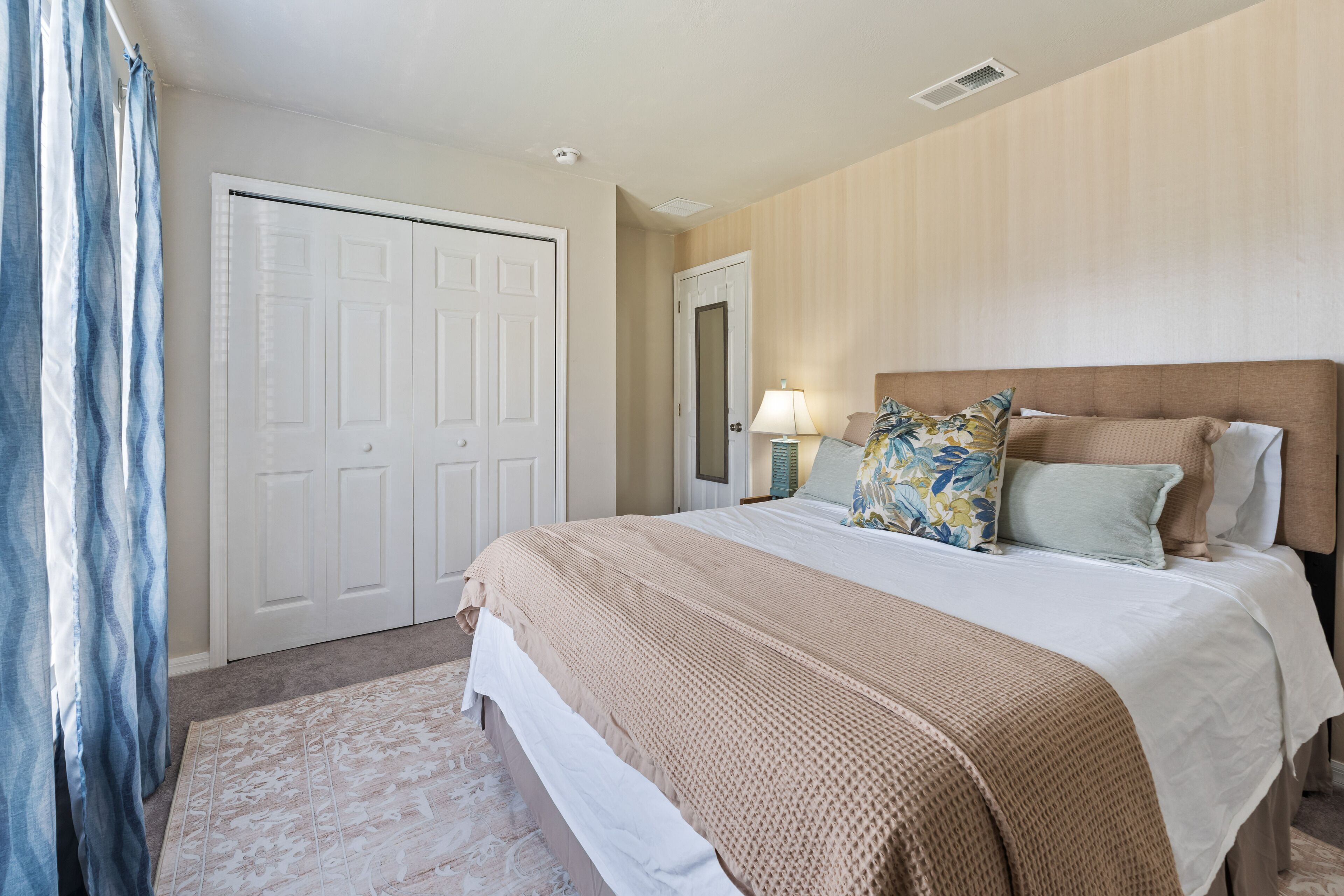 A bedroom with a large bed, a door, a nightstand with a lamp, and a window with curtains.