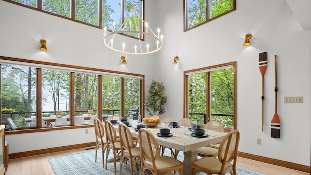 A dining area with a large table set for six, surrounded by wooden chairs, large windows, and a chandelier. - Family Luxury: 5BR, Beach, Kayaks, GameRm, Arcade, MovieRm, PingPong, Fire Pit (Saint Leonard)