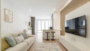 Een moderne woonkamer met een bank, twee barkrukken, een flatscreen-tv en een keuken in de achtergrond.