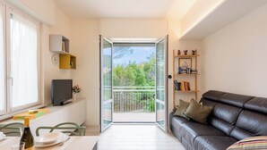 Un salon moderne comprenant un canapé, un téléviseur, une table à manger avec des chaises et un balcon offrant une vue sur la verdure.