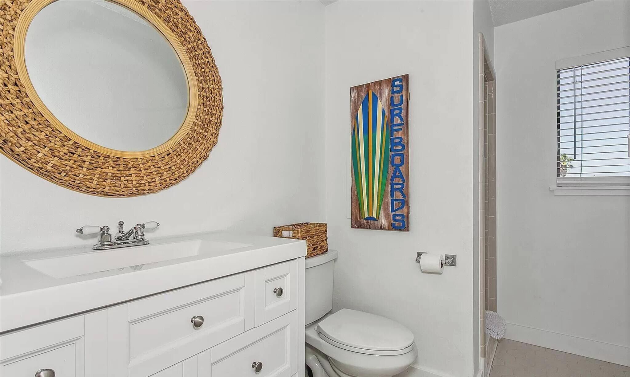 A bathroom with a white vanity, a round mirror with a woven frame, a toilet, and a shower area.