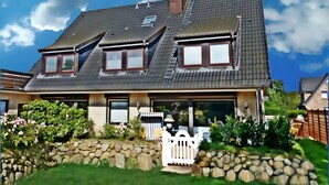 A two-story house with a sloped roof, a stone wall, and a white picket fence. - Beautiful Maisonette Apartment with Wi-Fi, Garden and Terrace; Parking Available (Sylt)