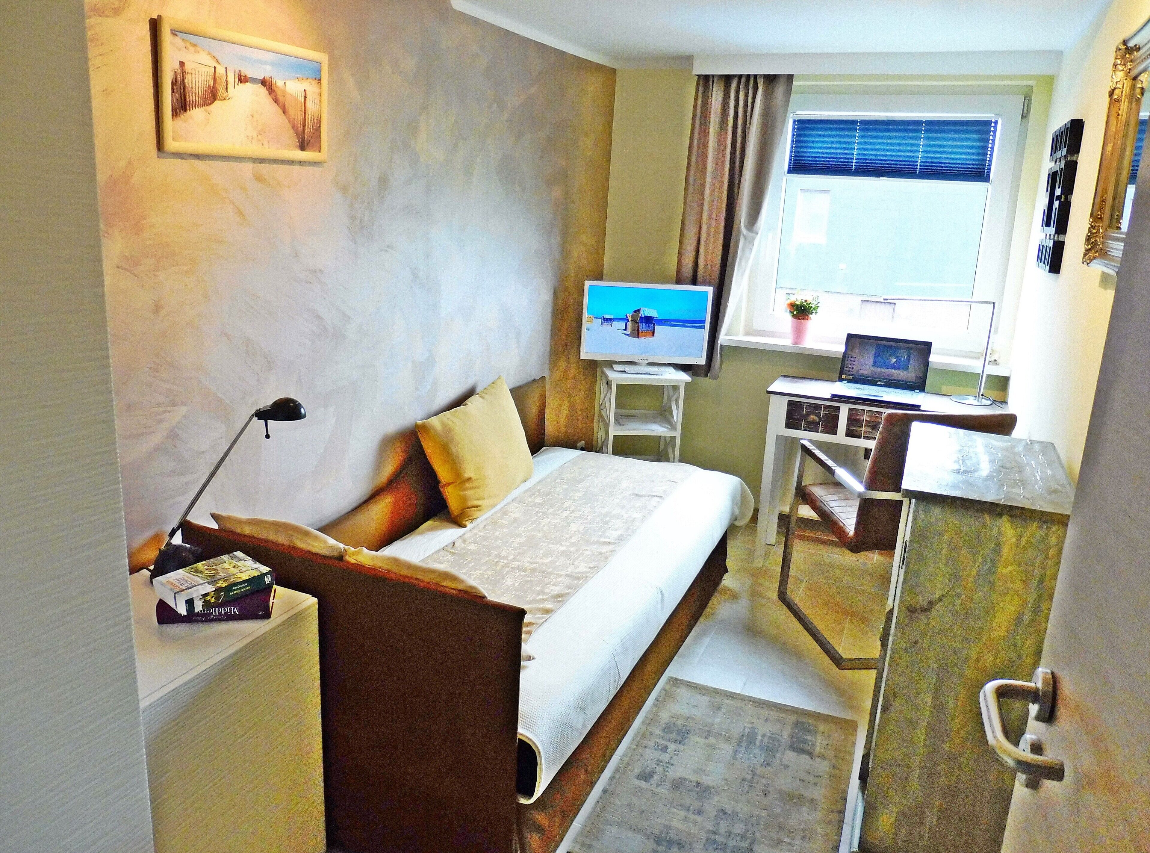Habitación de hotel compacta con cama, escritorio con una computadora portátil, televisor y ventana con cortinas.