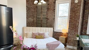 A modern bedroom with a brick wall, a large bed, a TV, and a vase of flowers. - Loft Apartment On Main - Sleeps 4 (Du Quoin)