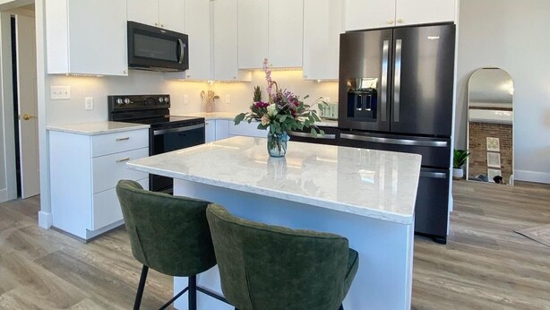 A modern kitchen with white cabinetry, a black refrigerator, a microwave, and a central island with two green bar stools. - Loft Apartment On Main - Sleeps 4 (Du Quoin)