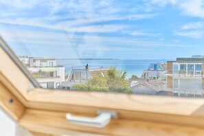 Blick aus einem Fenster auf eine Küstenlandschaft mit modernen Gebäuden und wolkenlosem Himmel.