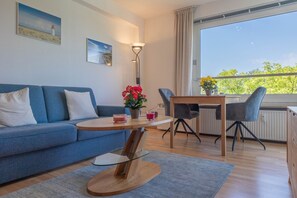 Uma sala de estar moderna com sofá, mesa de centro de vidro, mesa de jantar com cadeiras e vista para o verde através de uma janela.