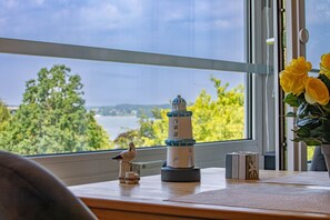 Une pièce avec vue sur une étendue d’eau, une maquette de phare et un vase rempli de fleurs jaunes posé sur une table.