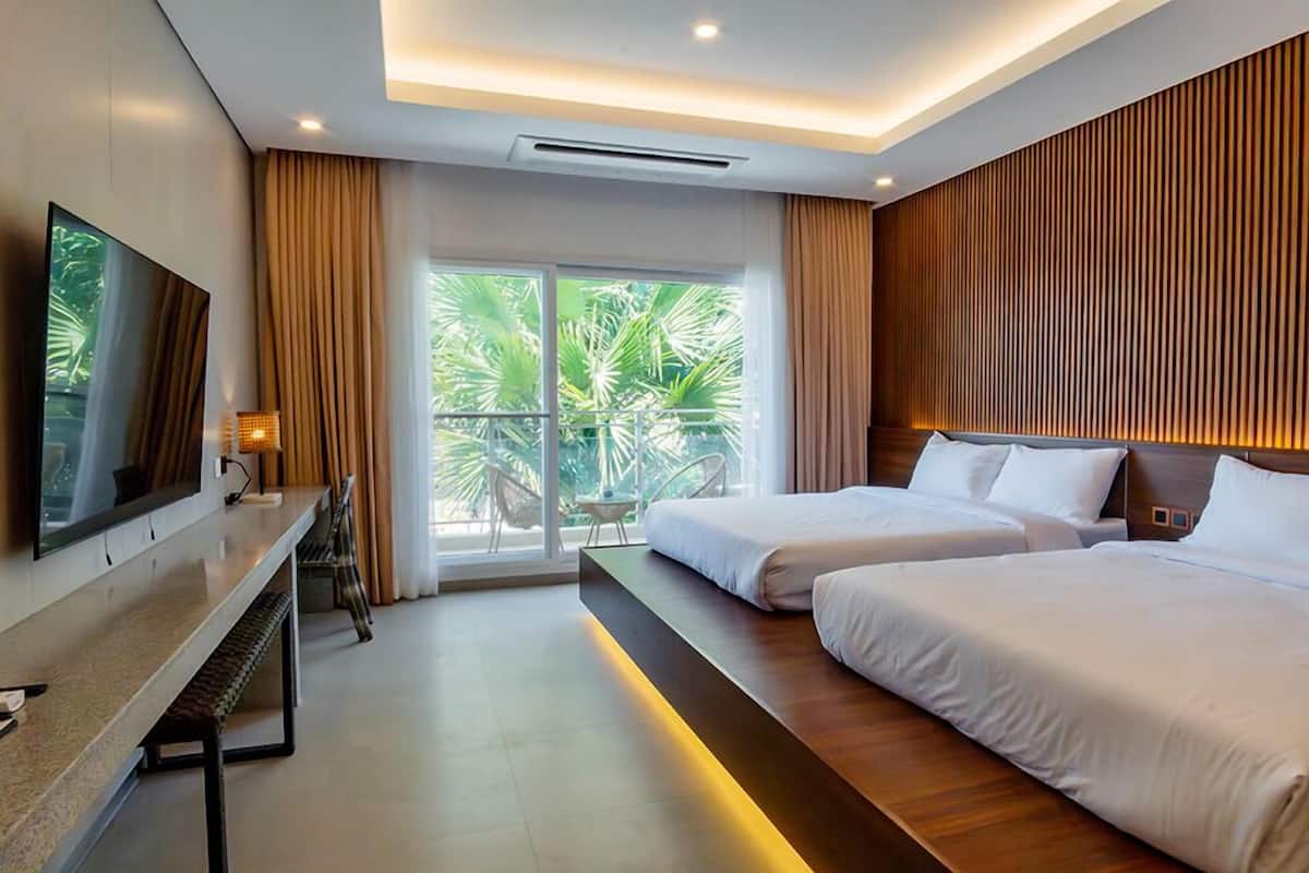 A modern hotel room with two beds, a flat-screen TV, a desk, and a balcony with a view of palm trees.