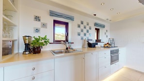 A modern kitchen with white cabinets, a stainless steel sink, and a built-in oven. There are decorative plates on the wall and a window with a view of the landscape. - Lütje Hüs 2 - Traumhafte Lage. Gemütlich. Natur pur (Hörnum (Sylt))