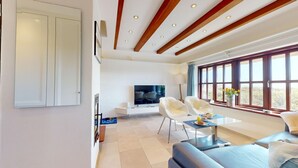 Un salon moderne avec un téléviseur, deux fauteuils blancs, un canapé et une table à manger où sont disposés des aliments pour le petit-déjeuner.