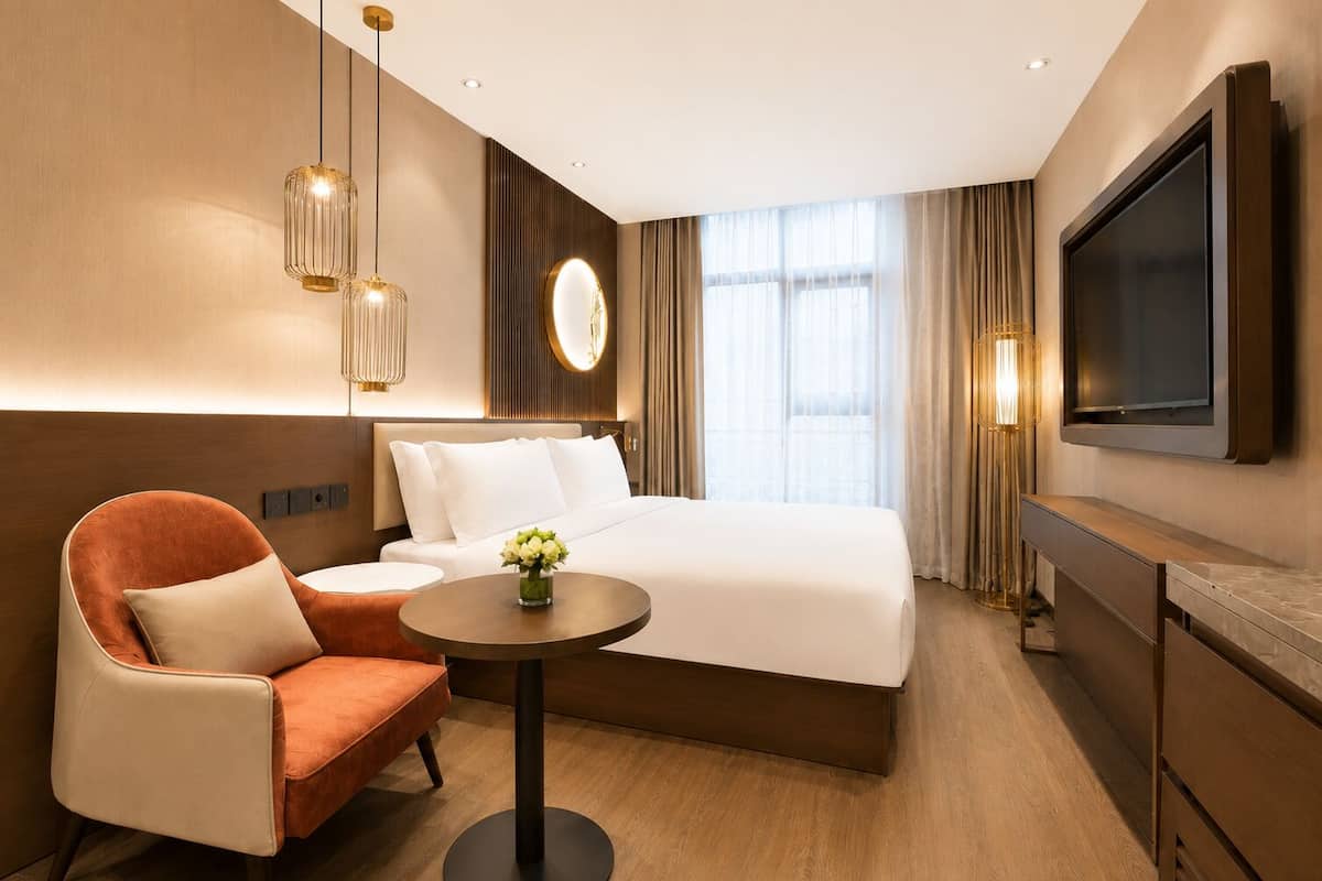 A hotel room with a bed, a round table with a vase, a chair, a TV, and a large window with curtains.