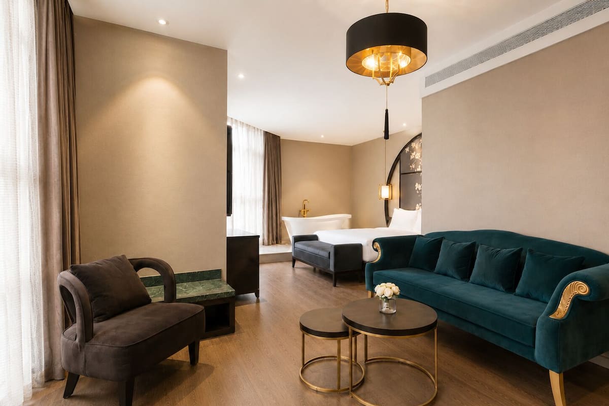A modern hotel room with a sofa, armchair, two small tables, a large window with curtains, a bathtub, and a chandelier.