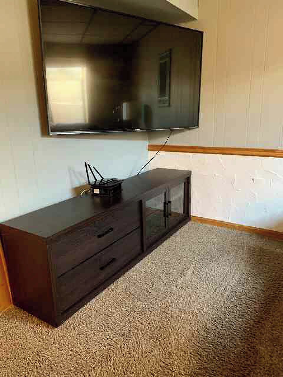 A room with a flat-screen TV mounted on the wall, a dark wooden cabinet below, and a beige carpet.