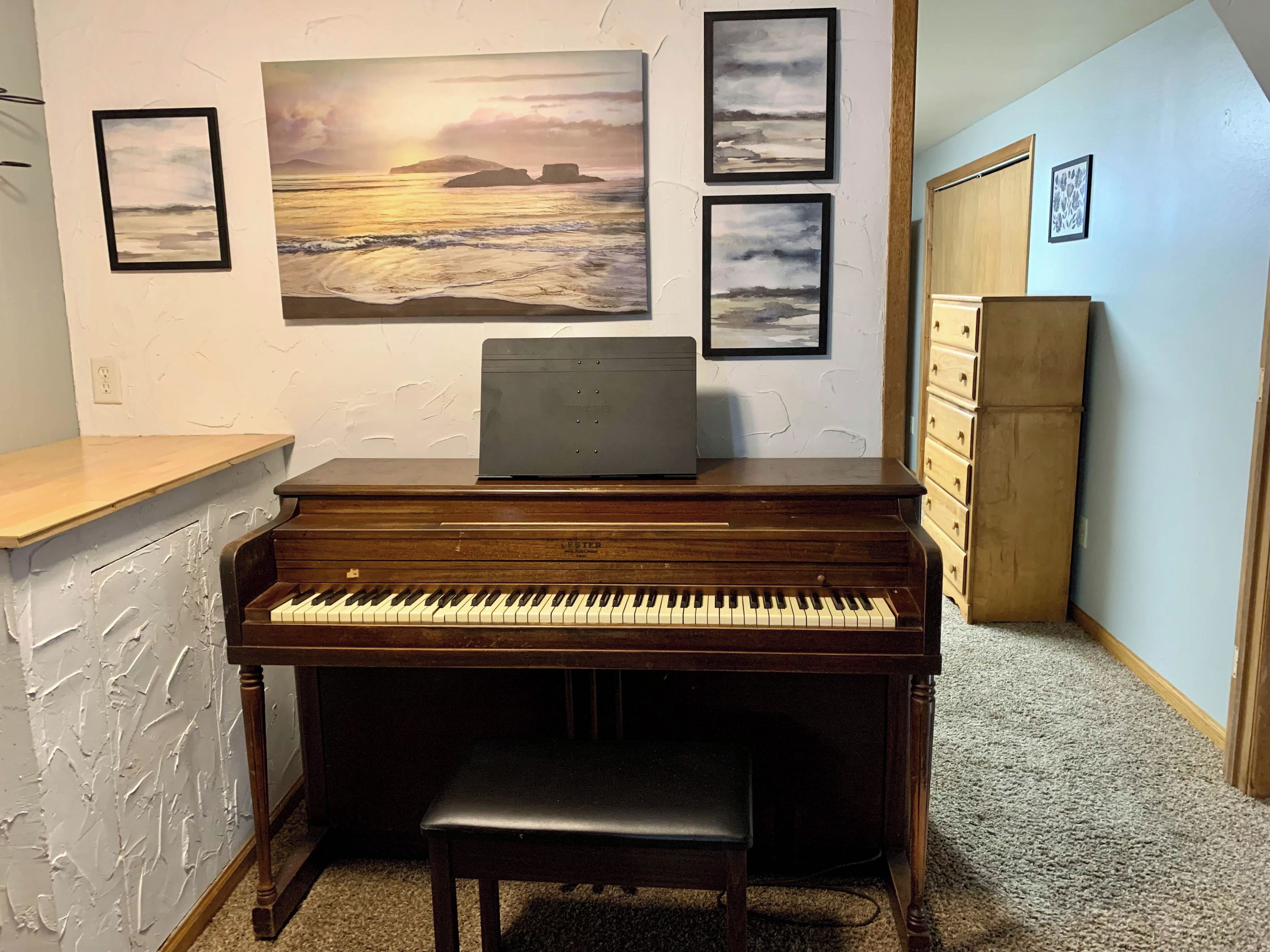A piano with a bench, a laptop on top, and framed artwork on the wall.