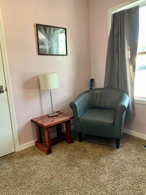 Quarto com poltrona cinza, mesa de cabeceira de madeira e luminária, quadro na parede e janela com cortina cinza.